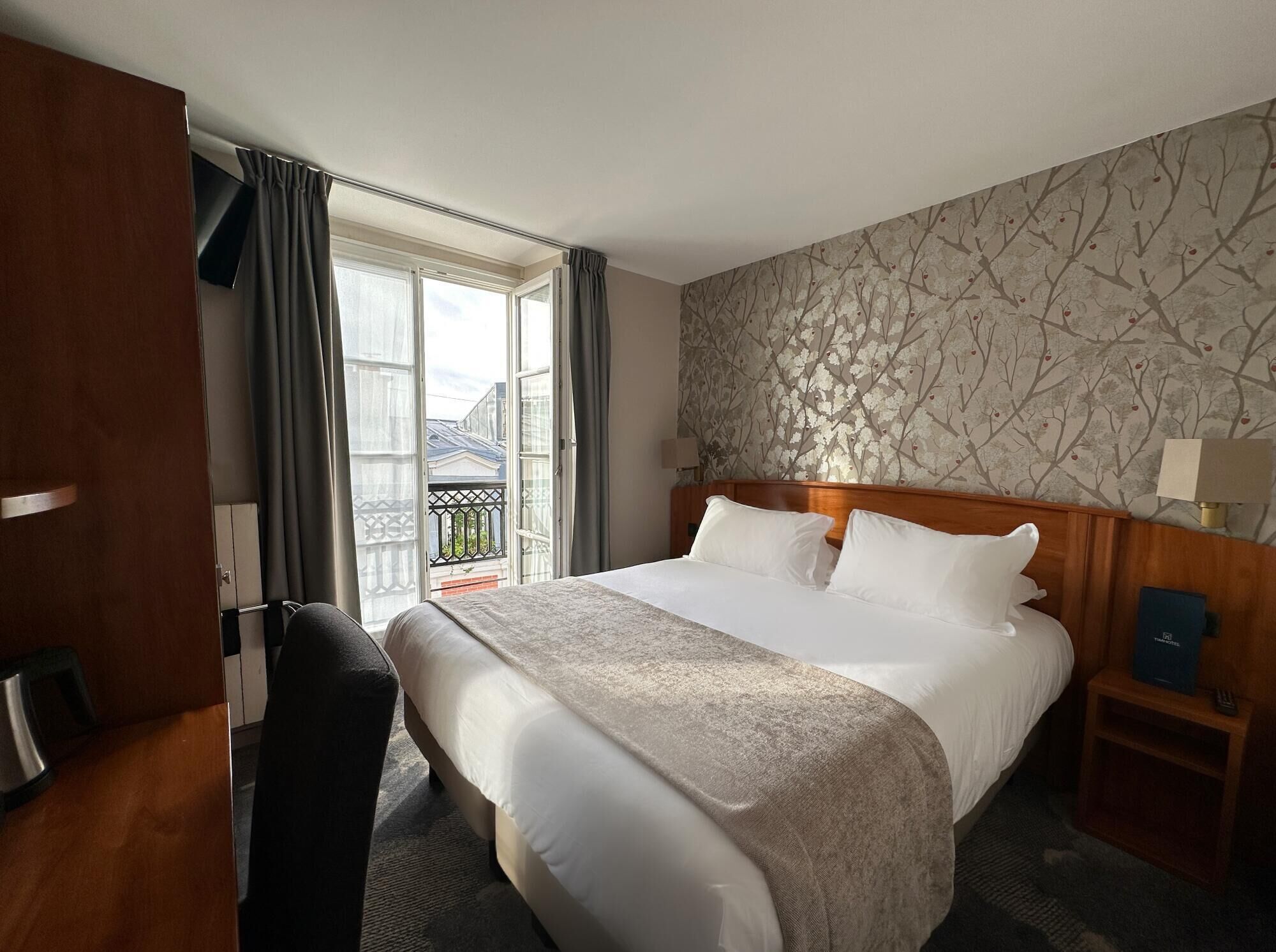Habitación de hotel con una cama grande, un cabecero de madera, una mesita de noche con lámpara y vistas al balcón exterior.
