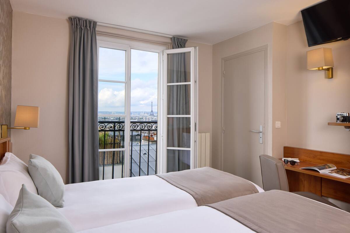 Une chambre d’hôtel avec deux lits, un bureau, une télévision et un balcon offrant une vue sur la Tour Eiffel.