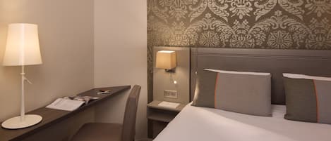 A hotel room with a desk, chair, lamp, and bed with pillows.