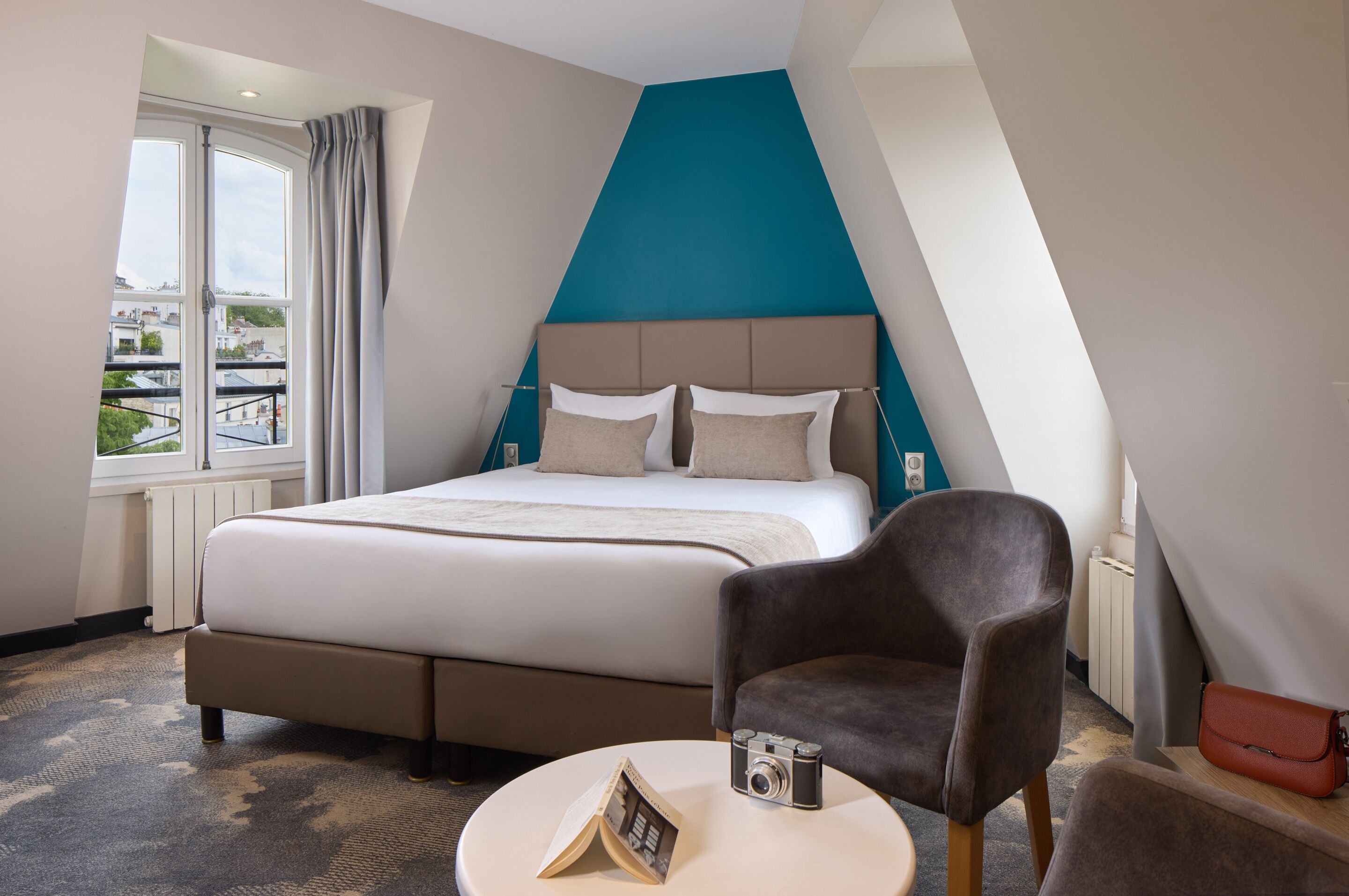 A hotel room with a large bed, a chair, a small table, and a view of buildings outside the window.
