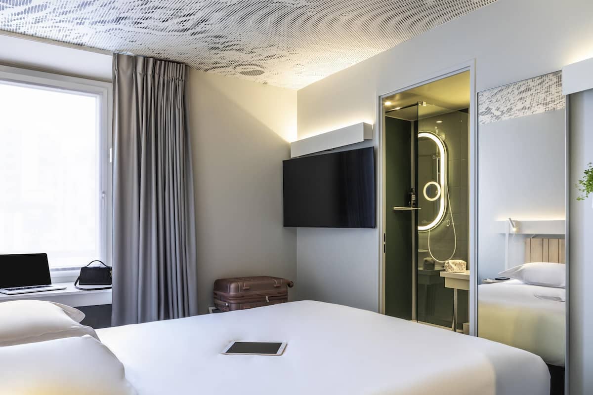 A modern hotel room with a bed, a desk with a laptop, a suitcase, and a bathroom with a glass door.