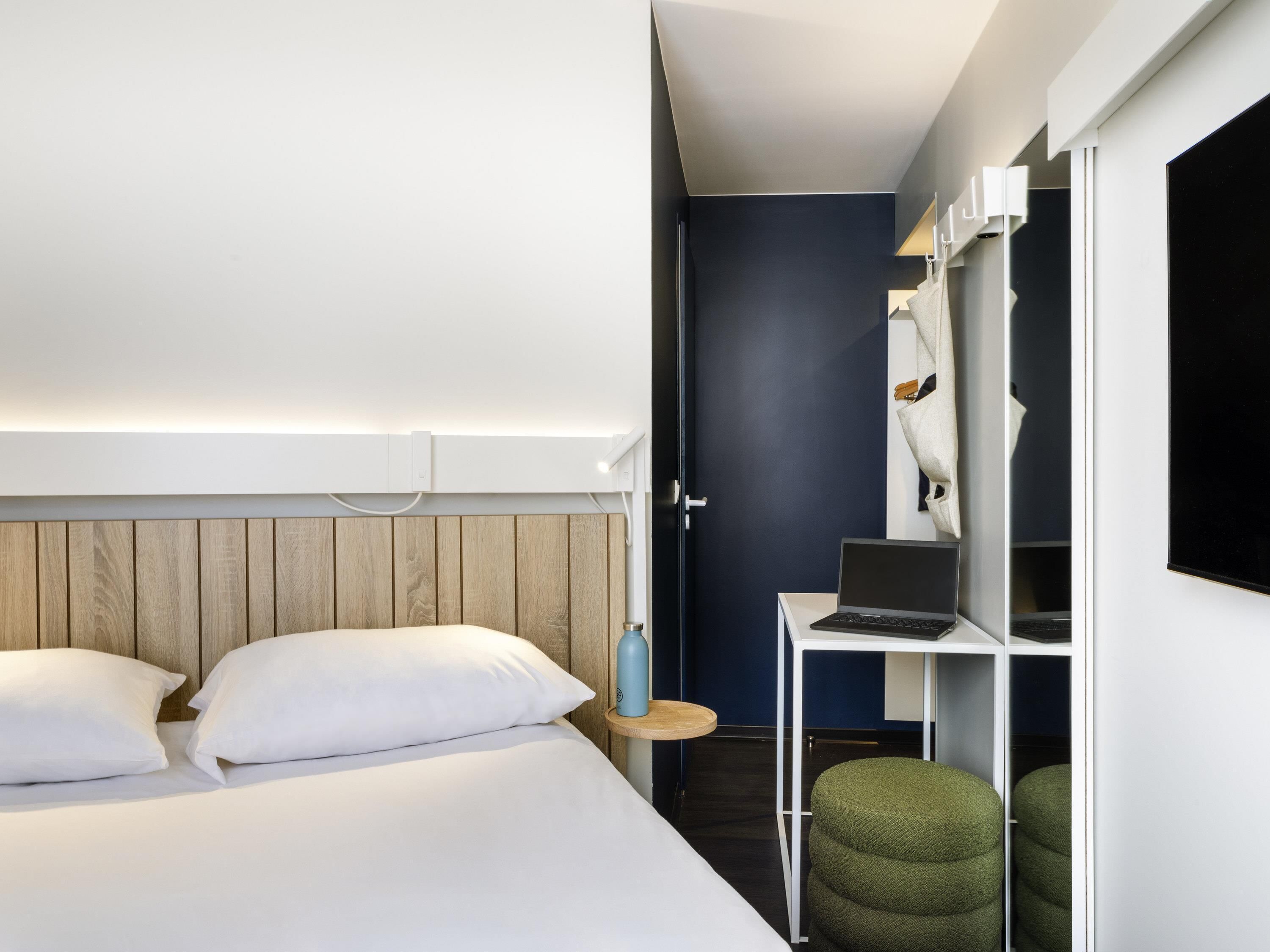 A modern hotel room with a bed, a desk with a laptop, and a wardrobe.