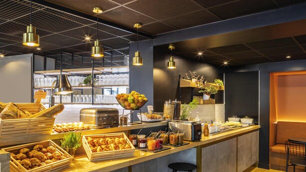 A modern café with a long counter, bar stools, hanging pendant lights, and a variety of food and drink options. - ibis Paris Nation Davout (Paris)
