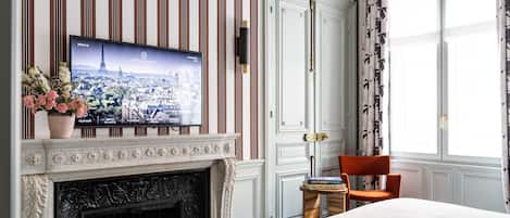 Habitación de hotel con televisión mostrando una vista urbana, chimenea, cama, silla y ventana con cortinas.