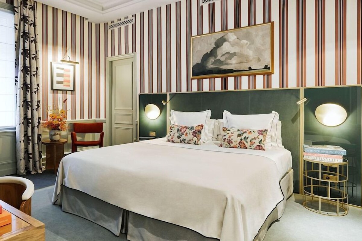 Quarto de hotel com uma cama grande, papel de parede listrado, um quadro e uma mesa de cabeceira com livros.