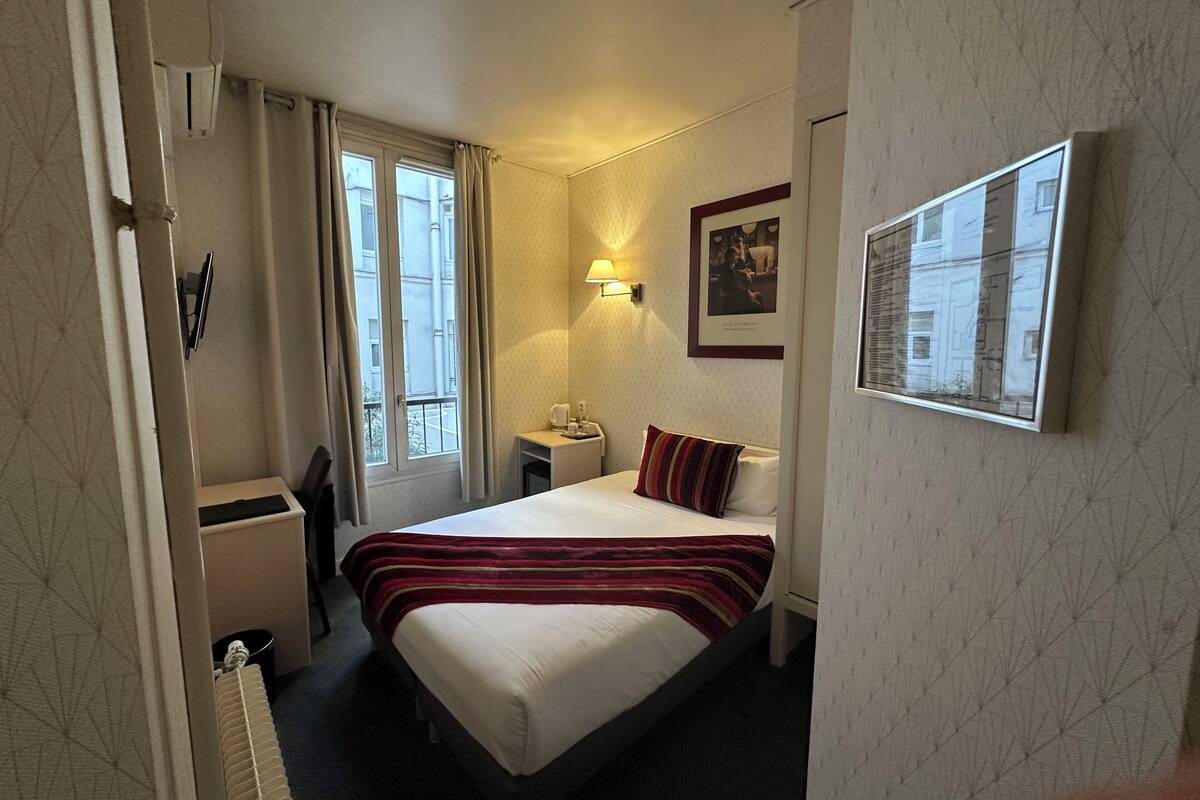 A hotel room with a bed, bedside table, lamp, and a window with curtains.