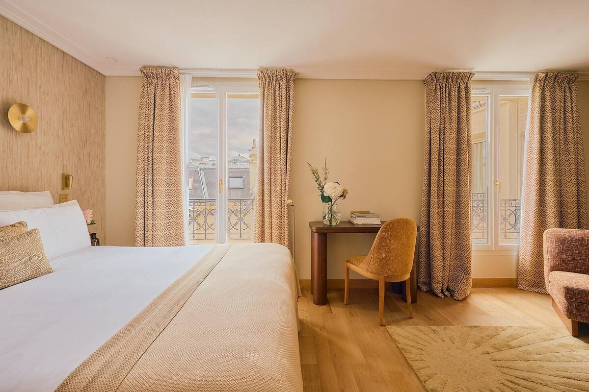 Une chambre d’hôtel avec un grand lit, un bureau avec une chaise et une vue sur la ville grâce à de grandes fenêtres ornées de rideaux à motifs.