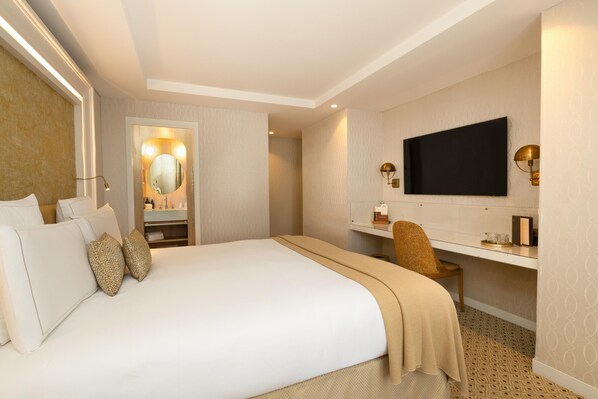 A hotel room with a large bed, a vanity with a mirror, a television mounted on the wall, and a desk with a chair. - Hôtel Madison (Paris)