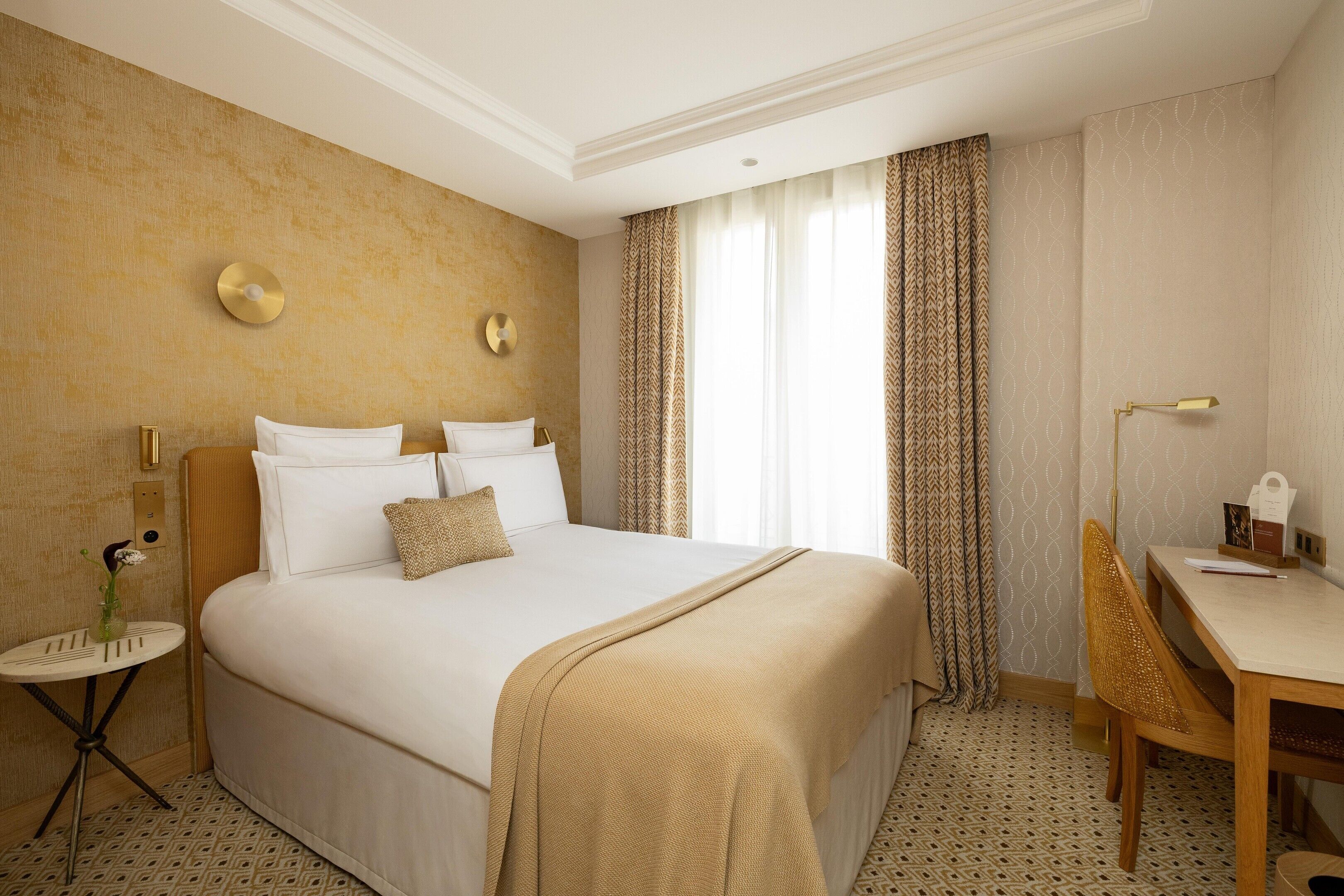 Una camera d'albergo con un letto grande, una scrivania e una sedia. Si vede una finestra con tende, una lampada a parete e un tavolino con un vaso.