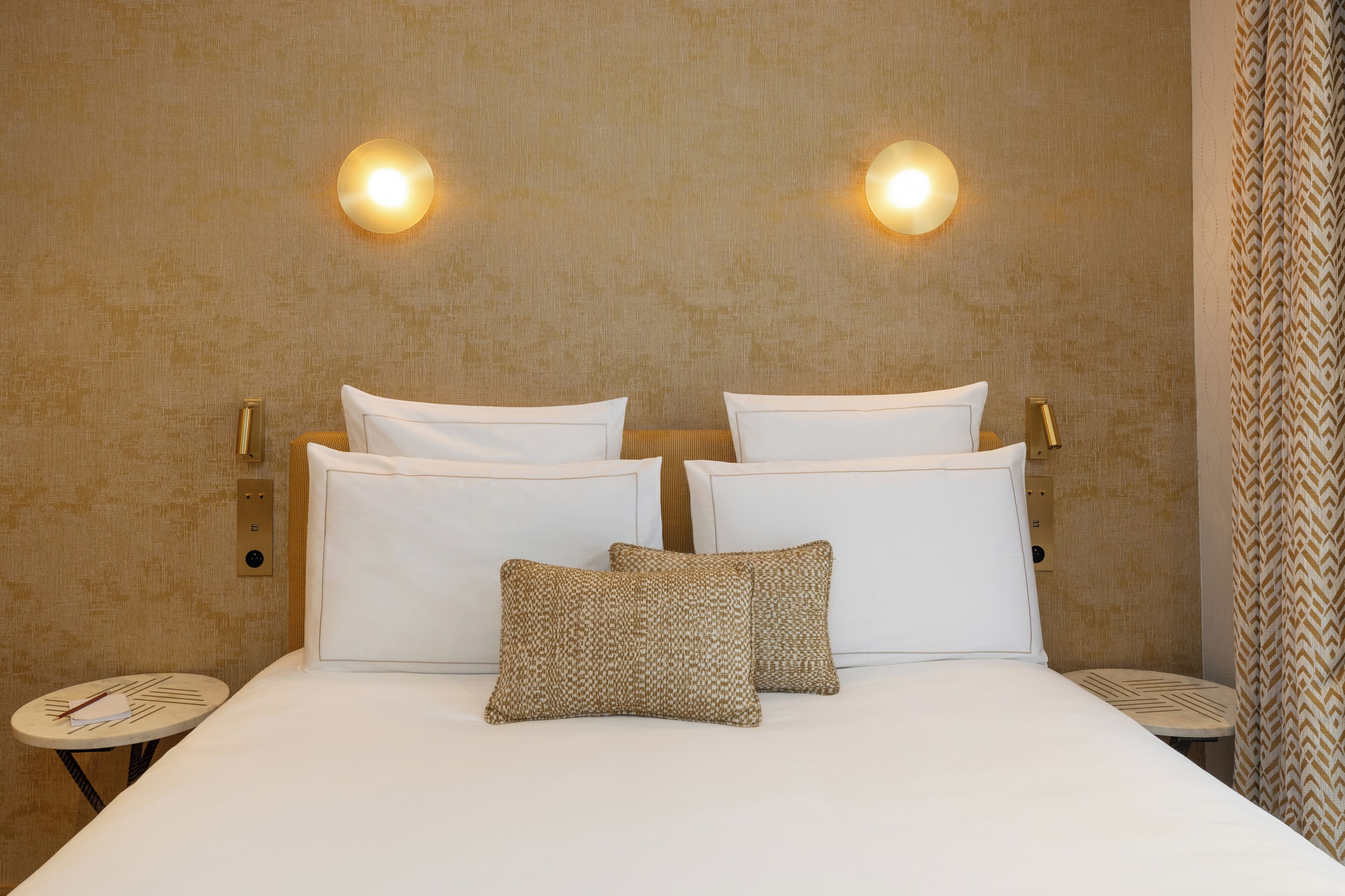 A hotel room with a bed, two wall-mounted lights, a bedside table, and a patterned throw pillow.