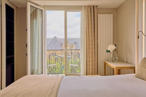 Une chambre d’hôtel avec un lit, une petite table sur laquelle est posé un vase de fleurs, une porte-fenêtre coulissante donnant sur un balcon aménagé avec du mobilier d’extérieur, et un rideau à motifs.