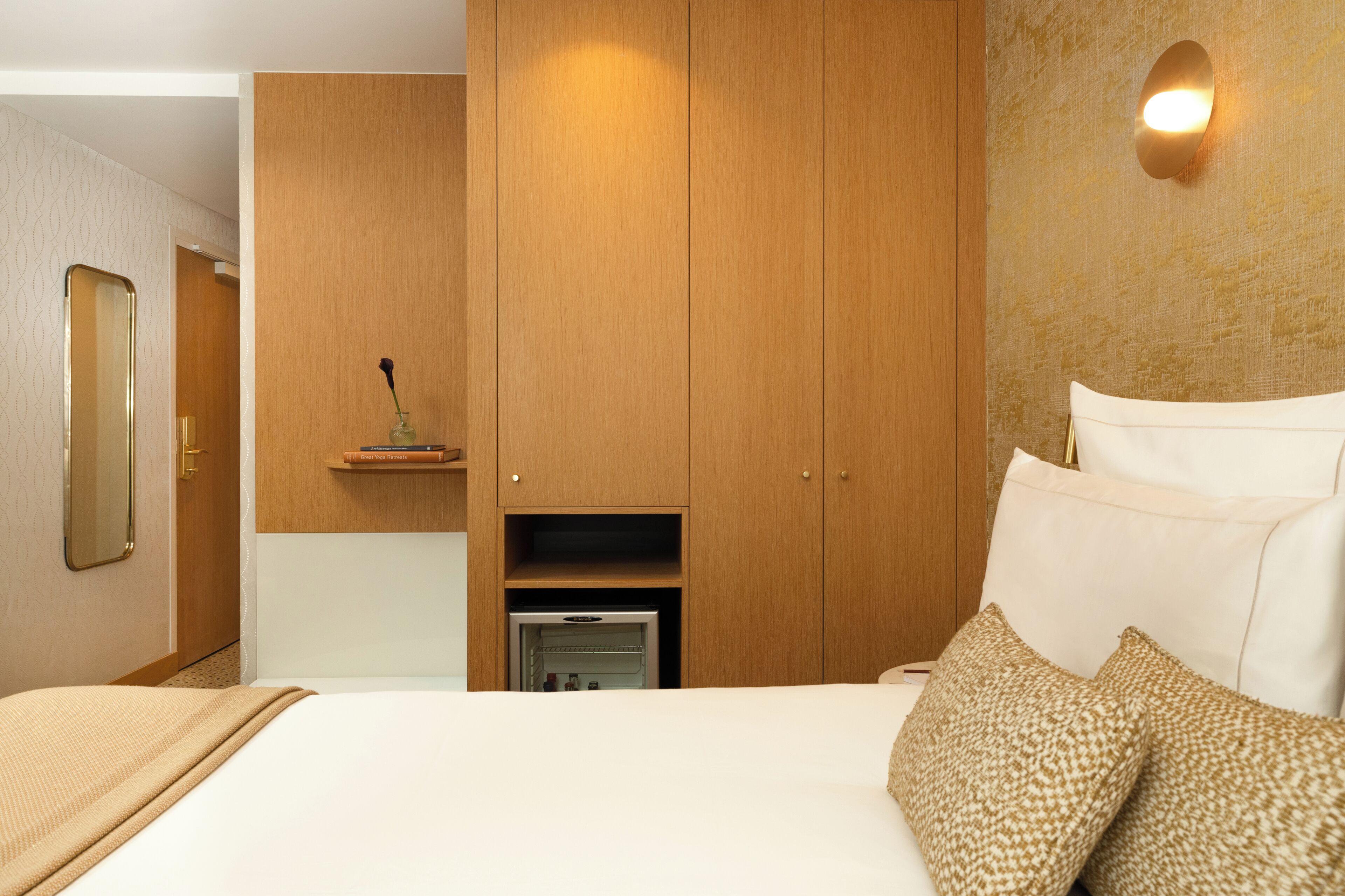 A hotel room with a bed, wooden wardrobe, a small shelf with a vase, and a wall-mounted light fixture.