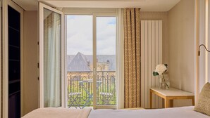 A hotel room with a bed, a small table with a vase of flowers, a sliding glass door leading to a balcony with outdoor furniture, and a patterned curtain. - Hôtel Madison (Paris)
