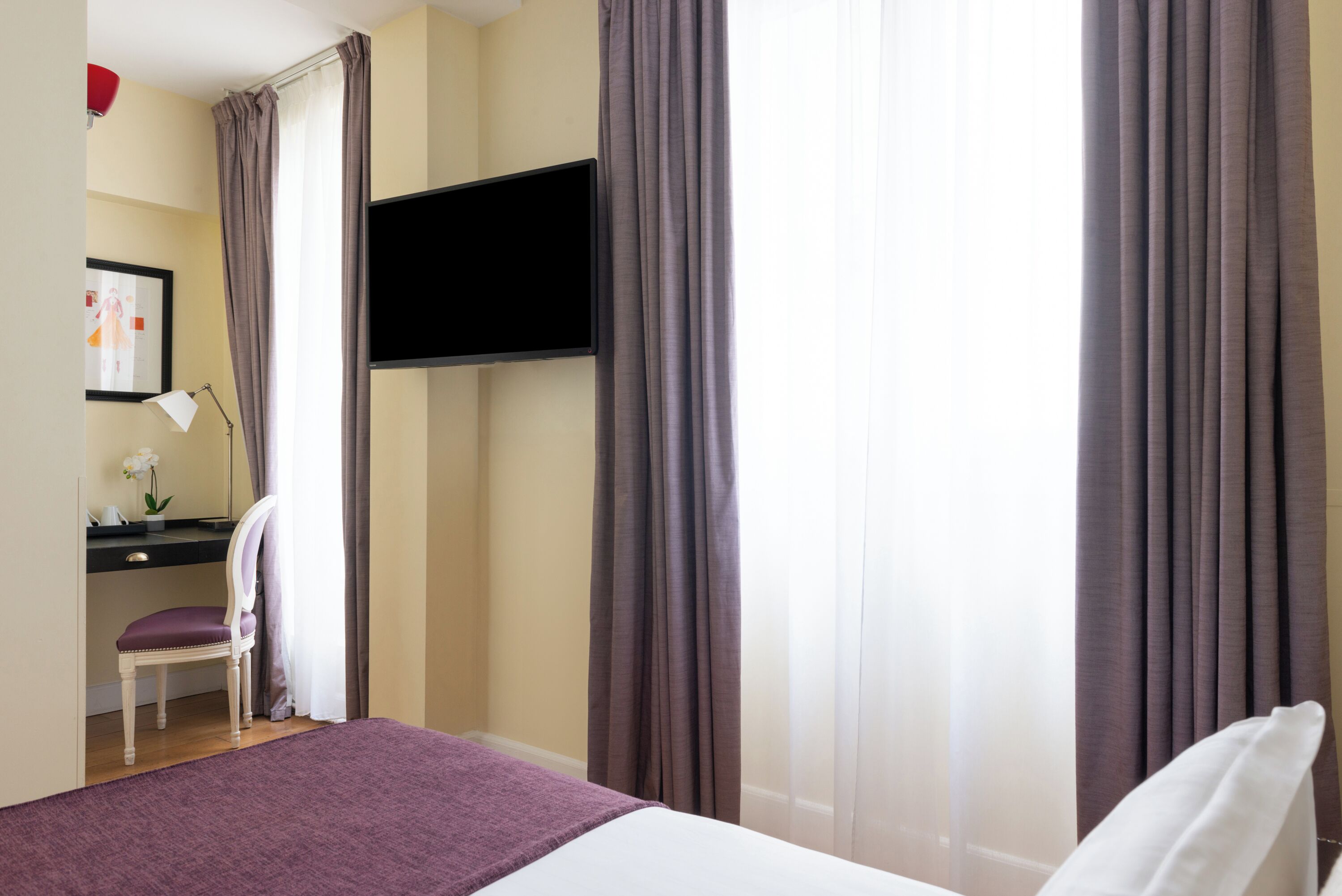 A hotel room with a bed, a desk with a chair, a TV mounted on the wall, and curtains.