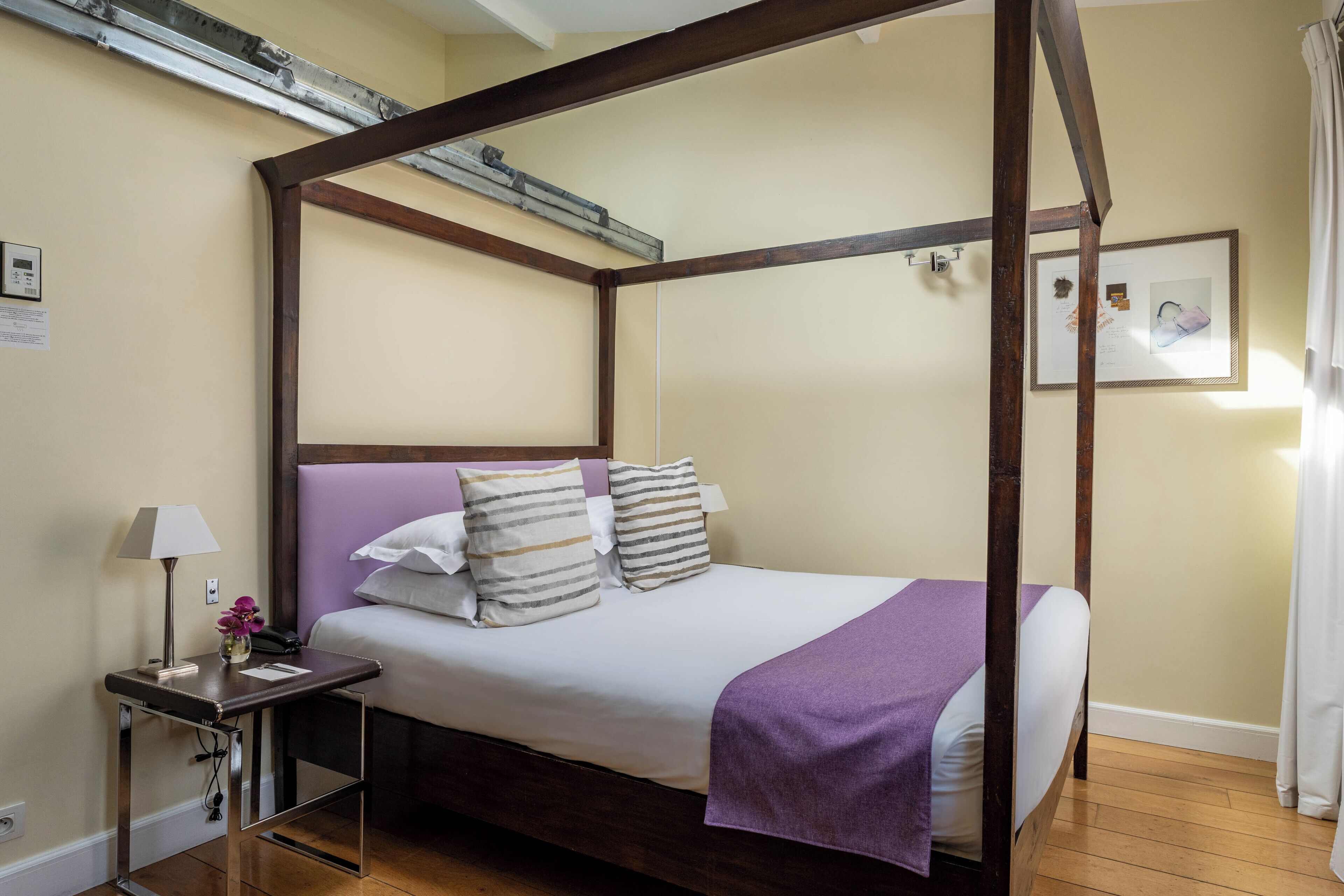 A four-poster bed with a wooden frame, a bedside table with a lamp, and a purple throw blanket.