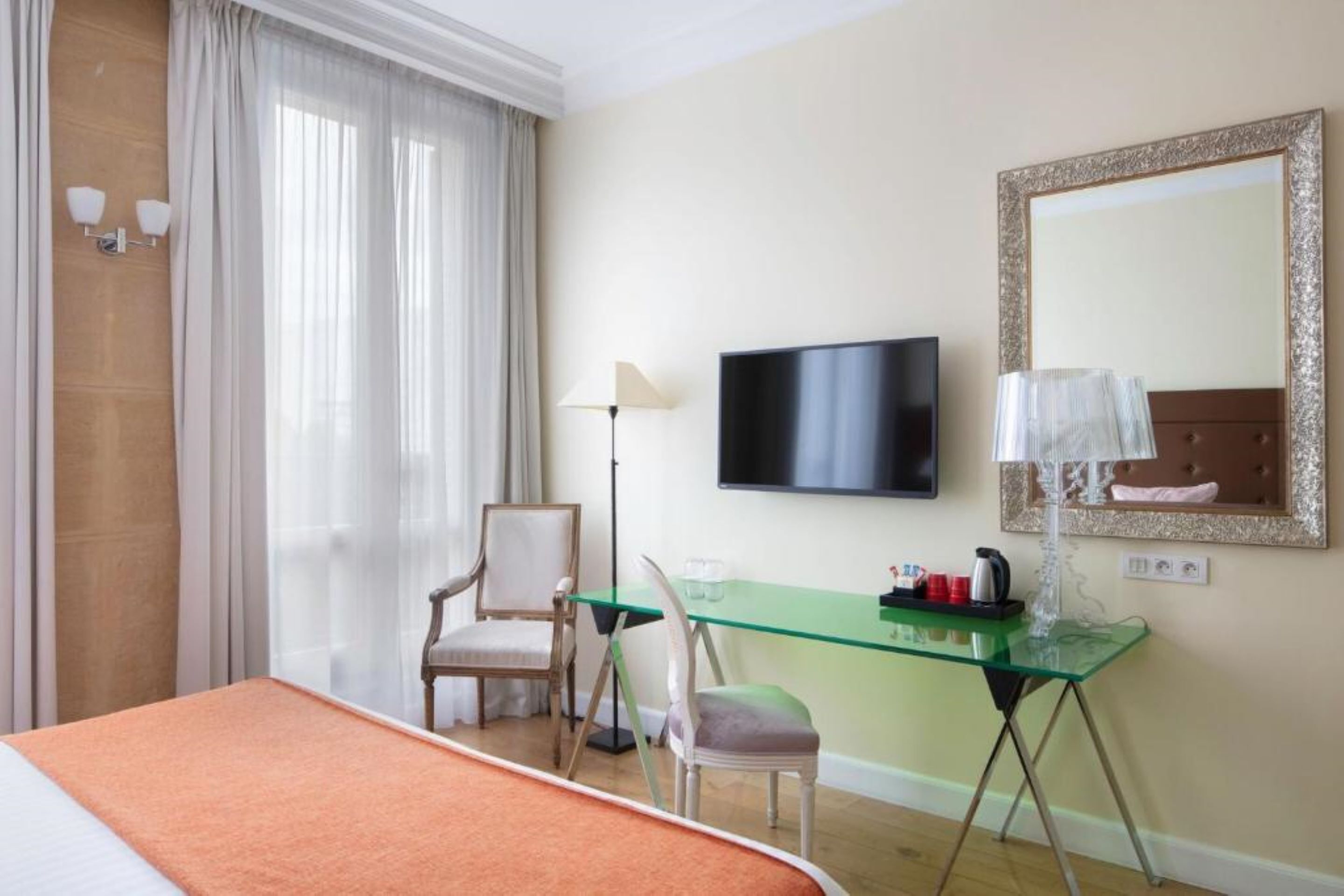 A hotel room with a bed, a desk with a chair, a television, a mirror, and a window with curtains.