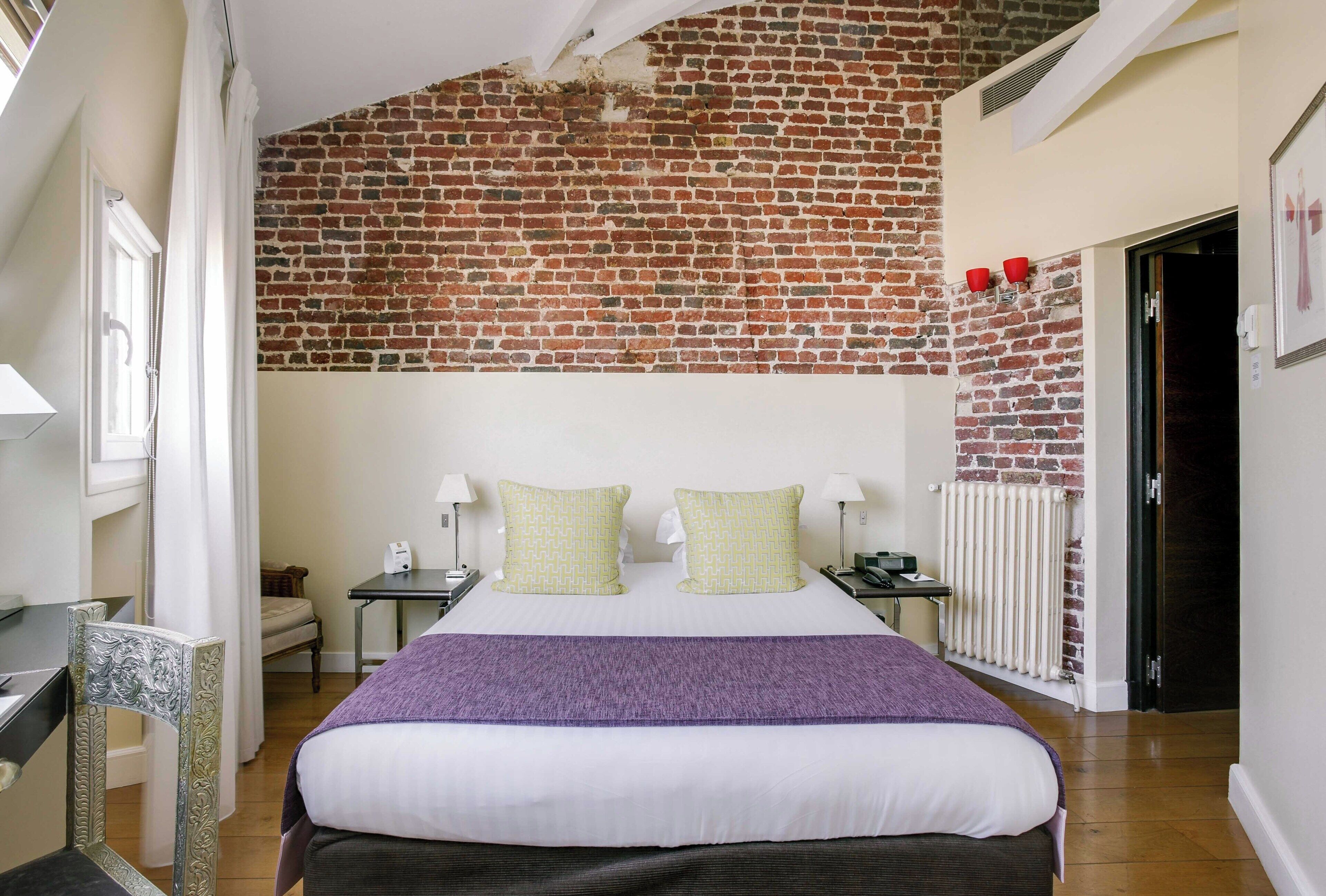 Een hotelkamer met een bakstenen muur, een bed met wit en paars beddengoed, twee nachtkastjes met lampen en een stoel.
