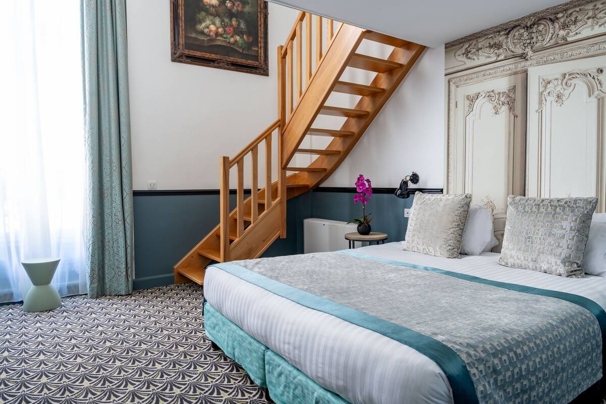 A bedroom with a bed, a wooden staircase, a small table with a vase, and a wall-mounted painting.