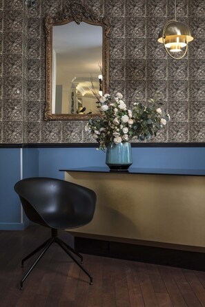 A reception area with a dark wooden floor, a black chair, a blue vase with white flowers, a mirror with an ornate frame, and a wall with a floral pattern.