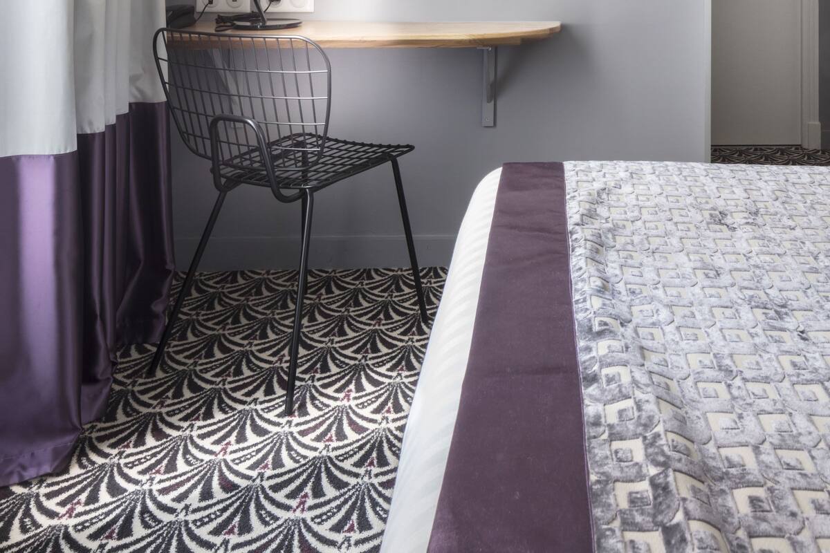 A hotel room with a bed, a desk with a chair, a mirror, and a wall-mounted lamp.