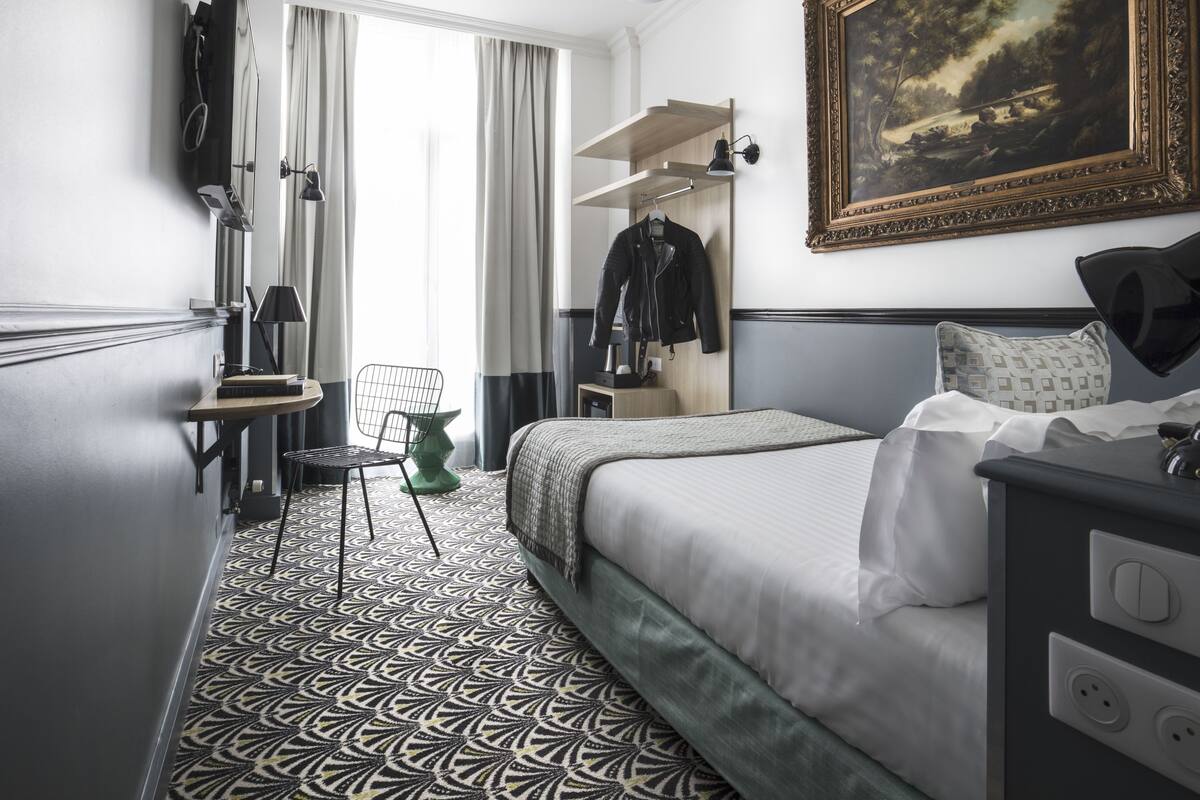 A hotel room with a bed, a desk, a chair, a coat rack, a painting, and a patterned carpet.