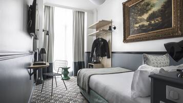 Habitación de hotel con cama, escritorio, silla, perchero, un cuadro y alfombra estampada.