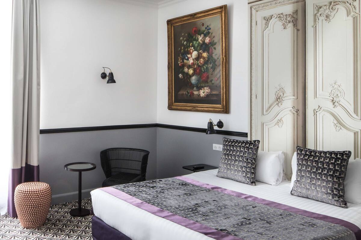 A hotel room with a bed, a small round table, a chair, a decorative painting, and a door with ornate details.