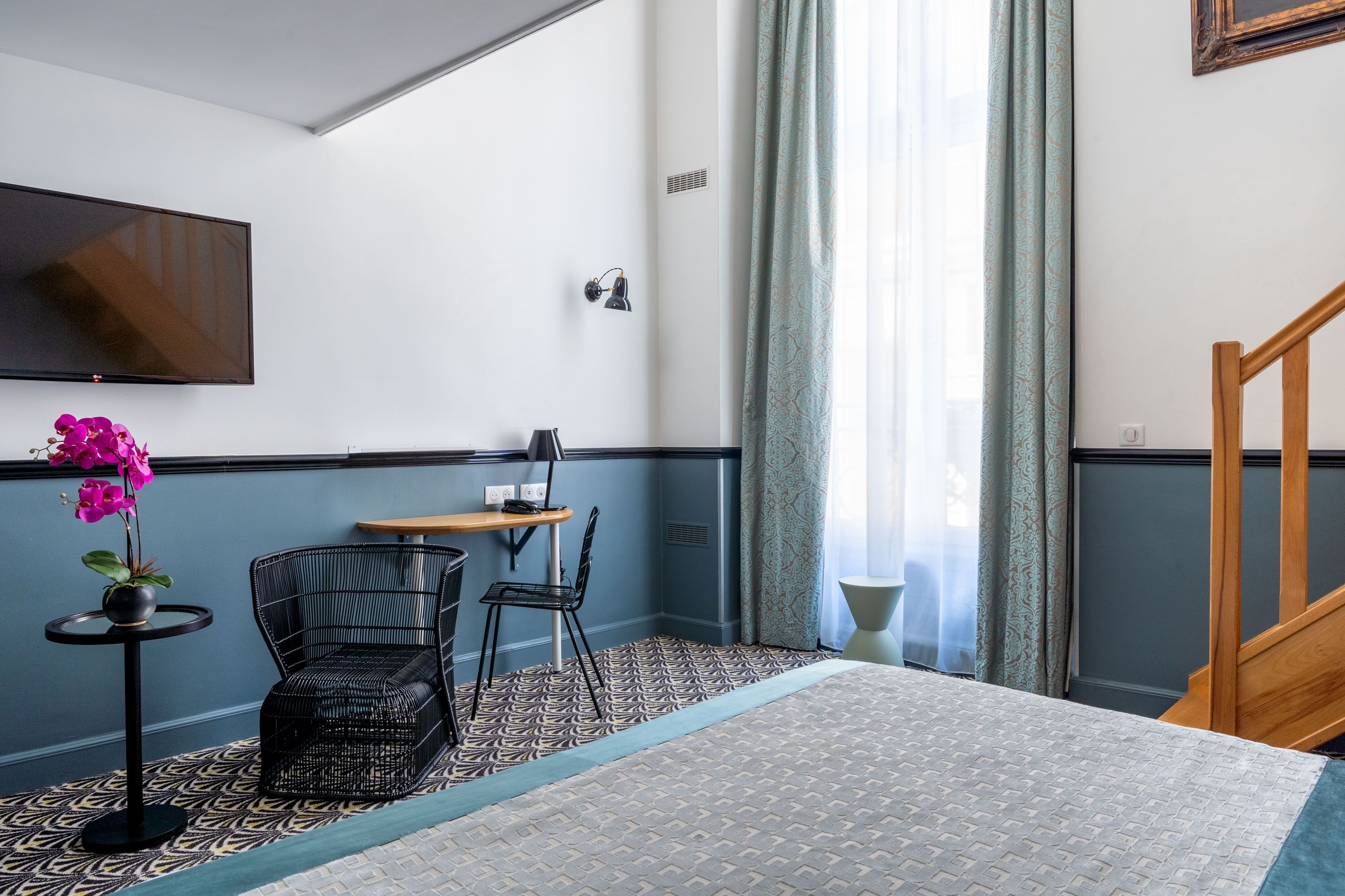 A hotel room with a bed, a TV mounted on the wall, a small table with a chair, a potted plant, and a patterned rug.