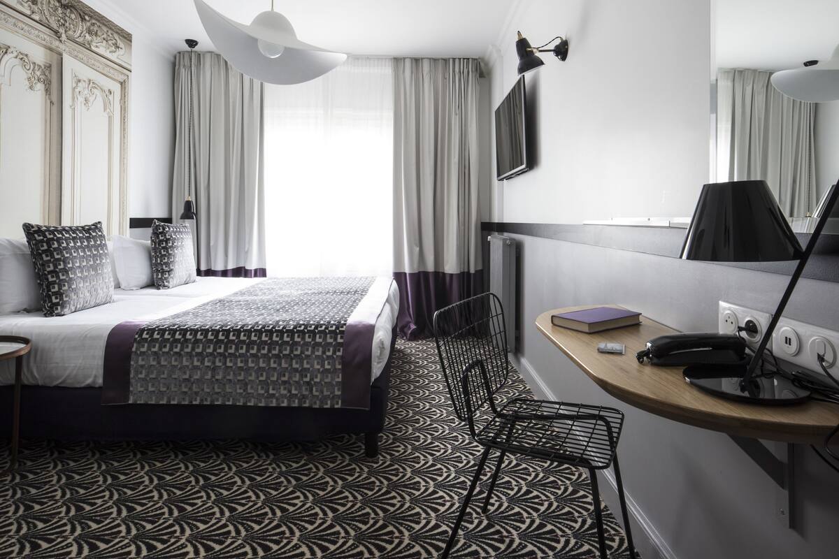 A modern hotel room with a large bed, a desk with a lamp, a chair, and a large mirror.