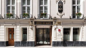 La façade d’un hôtel, avec une enseigne indiquant « HÔTEL DE MALTE », de grandes fenêtres et des jardinières remplies de fleurs sur les balcons.