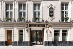 La façade d’un hôtel, avec une enseigne indiquant « HÔTEL DE MALTE », de grandes fenêtres et des jardinières remplies de fleurs sur les balcons.