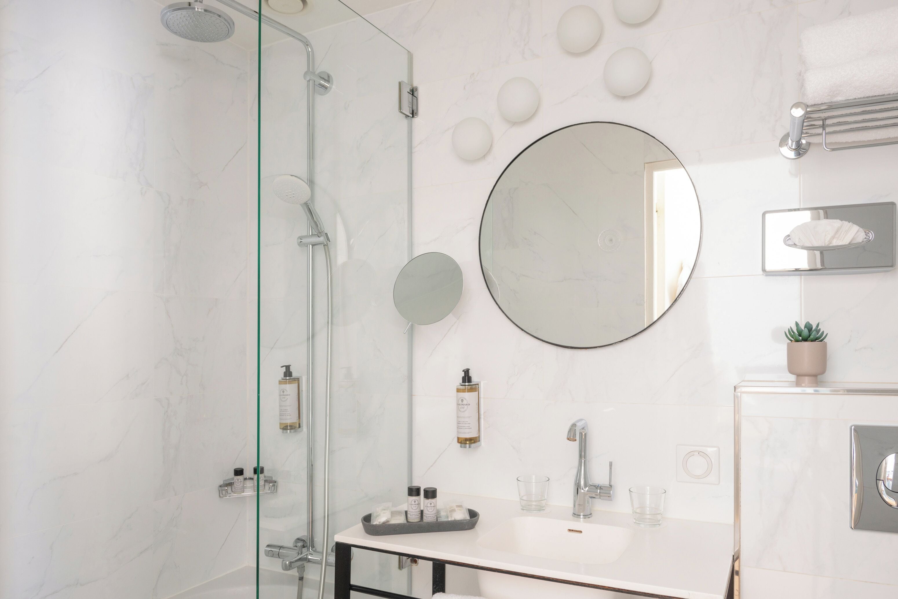 A modern bathroom with a glass-enclosed shower, a round mirror, a white sink, and a small potted plant.