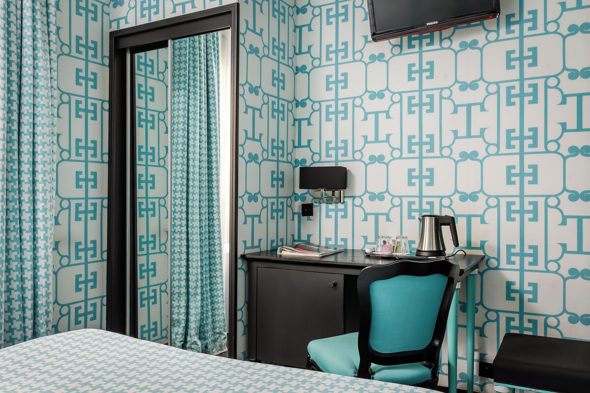 A hotel room with a patterned wallpaper, a desk with a lamp, a chair, and a television on the wall.
