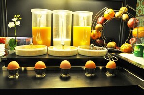 A kitchen counter with four glass containers of orange juice, a fruit wire rack with oranges and apples, and several egg holders. - Hôtel Monceau Elysées (Paris)