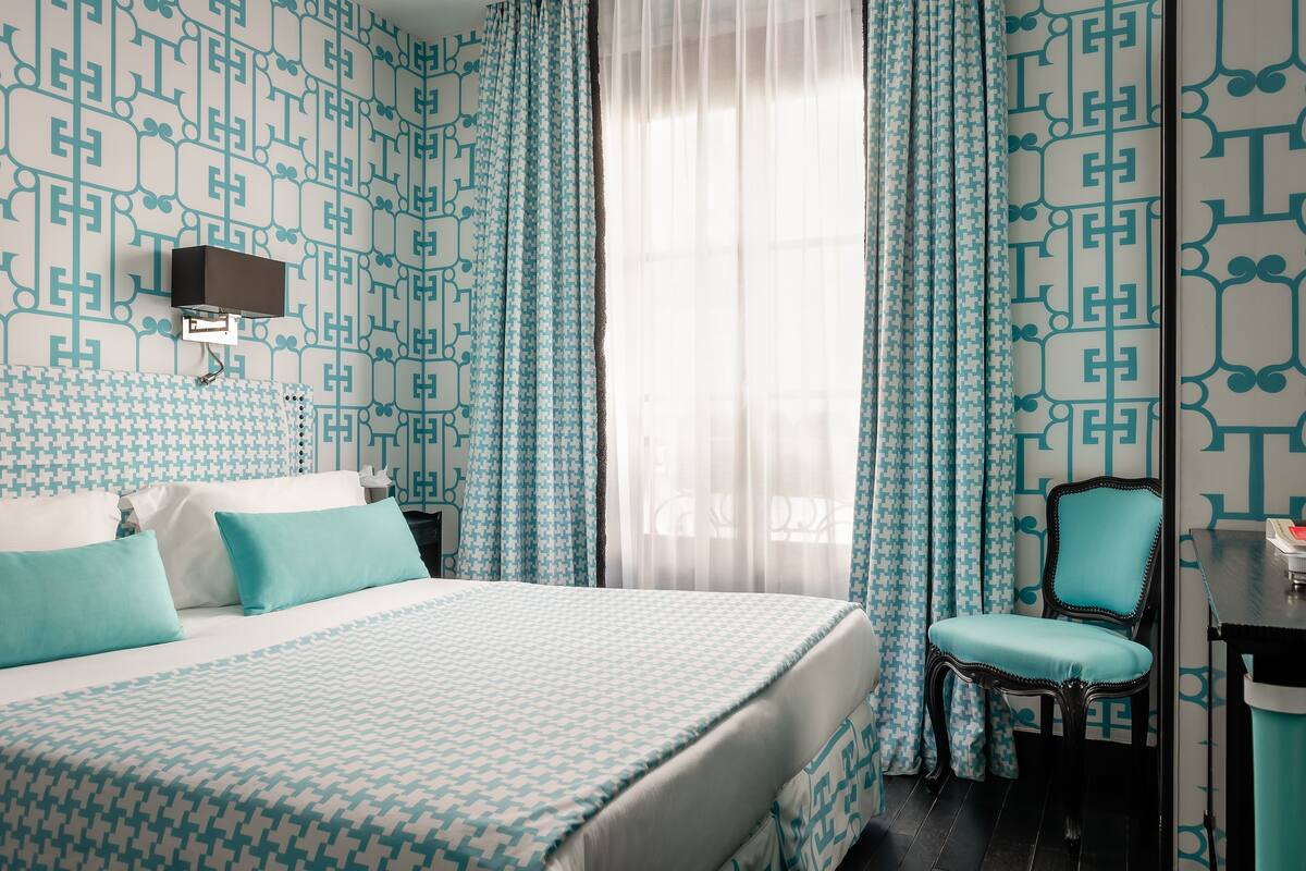 A hotel room with a patterned wallpaper, a bed with a patterned bedspread, a chair, a desk, and a television on the wall.