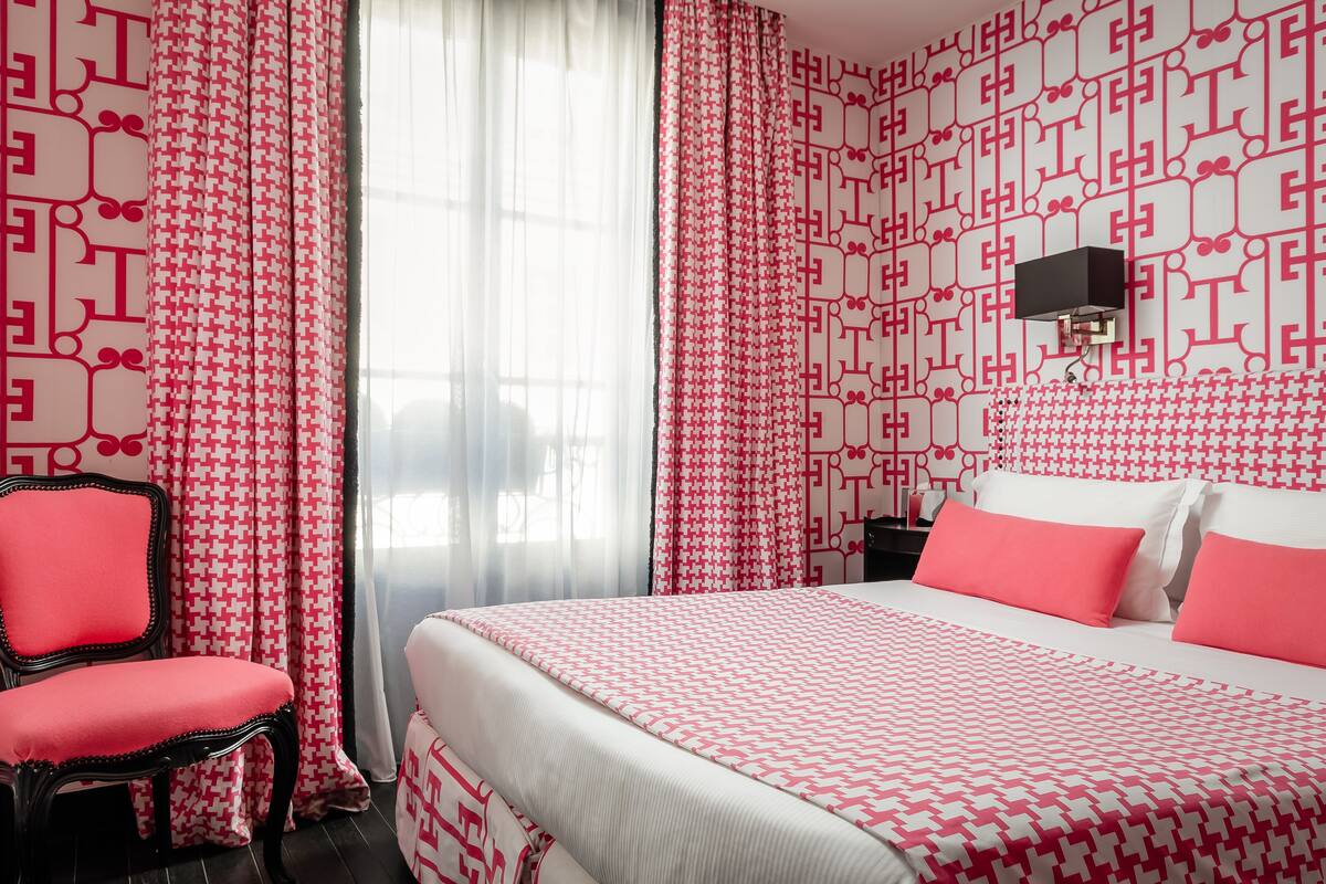 A hotel room with a patterned wallpaper, a bed with pink and white bedding, a chair, and a wall-mounted TV.