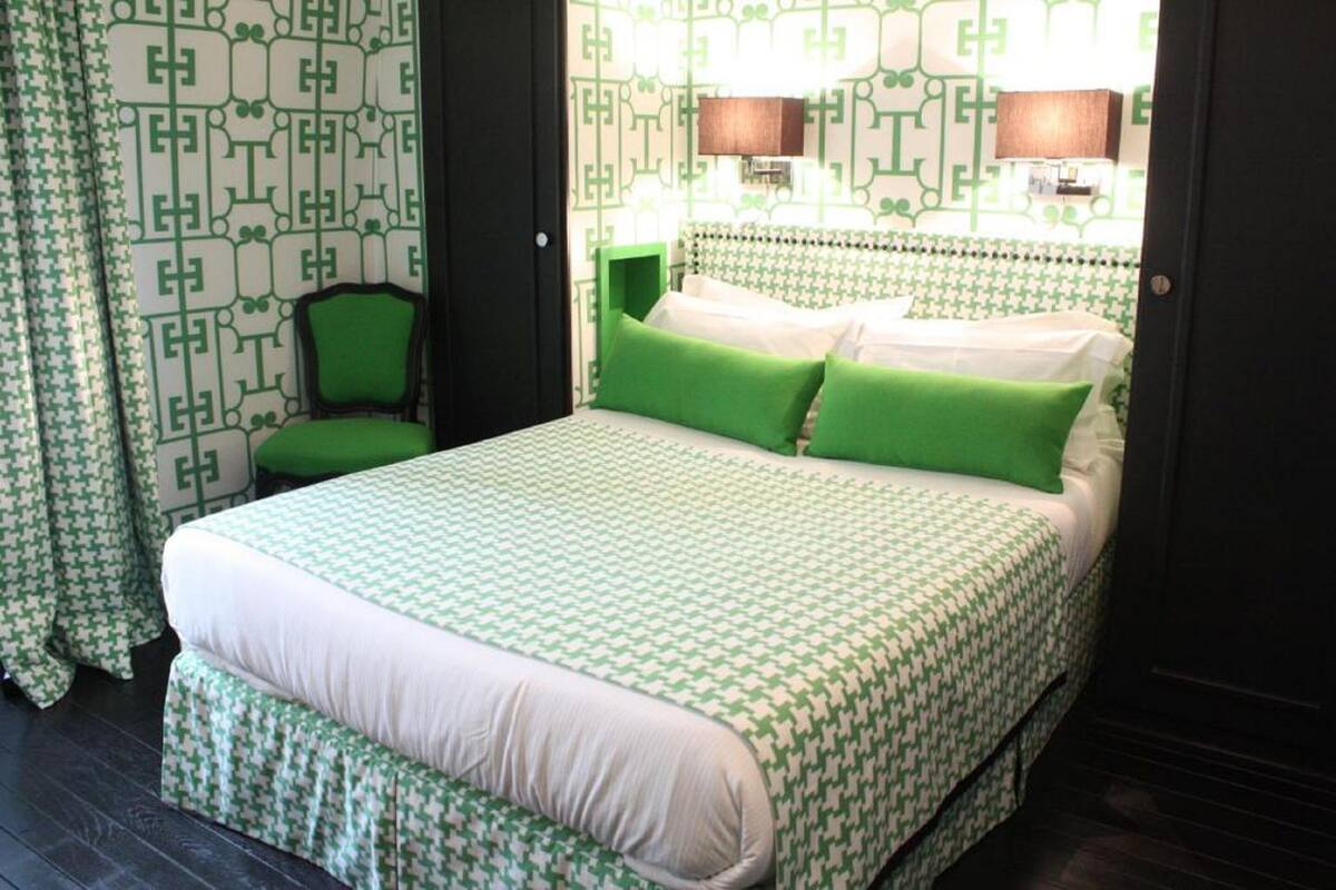 A hotel room with a bed, green and white patterned wallpaper, two bedside lamps, and a green chair.
