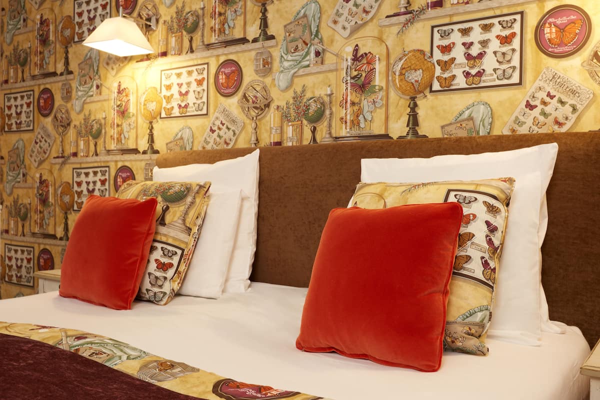 Una cama con cabecera marrón, ropa de cama blanca y dos almohadas rojas. La pared detrás de la cama está decorada con arte con temática de mariposas.