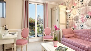 Quarto com escrivaninha, duas cadeiras, sofá e vista para um edifício pela janela.