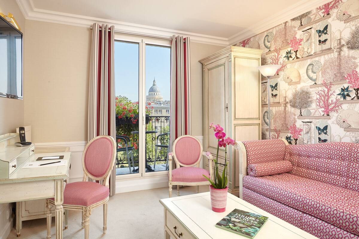 Una habitación con escritorio, dos sillas, un sofá y vistas a un edificio a través de una ventana.