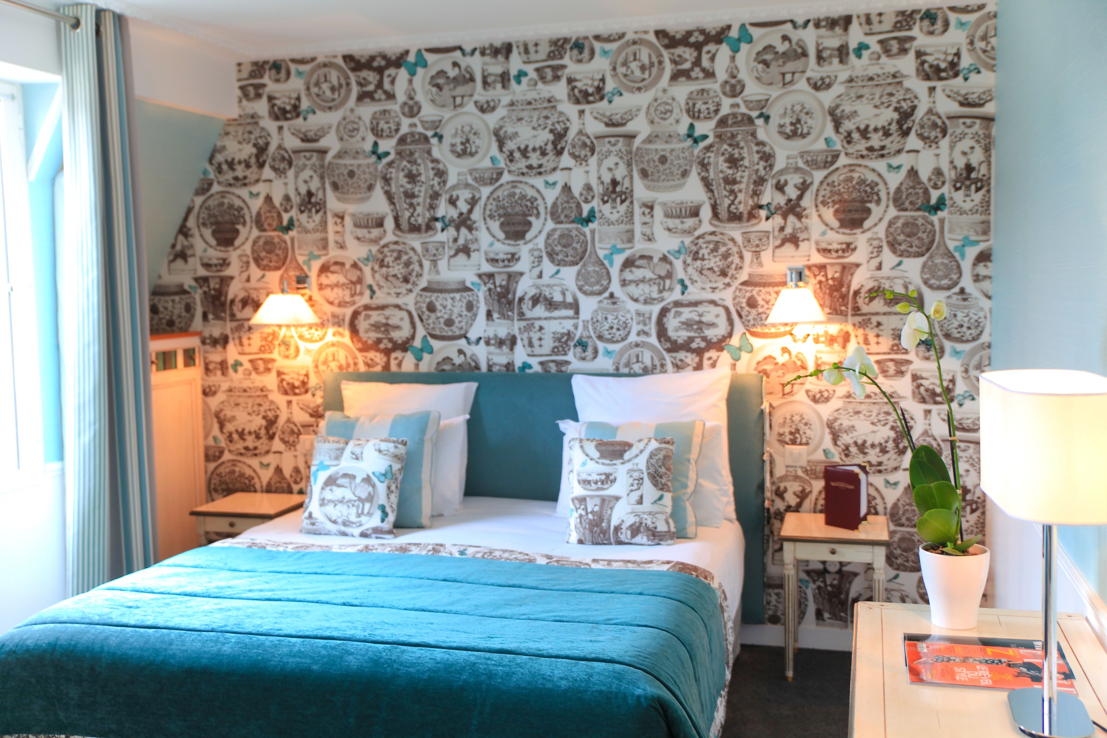 Une chambre d’hôtel avec un papier peint à motifs, un lit avec une literie de couleur turquoise, deux tables de chevet avec des lampes et une petite table sur laquelle se trouvent une plante et un livre.