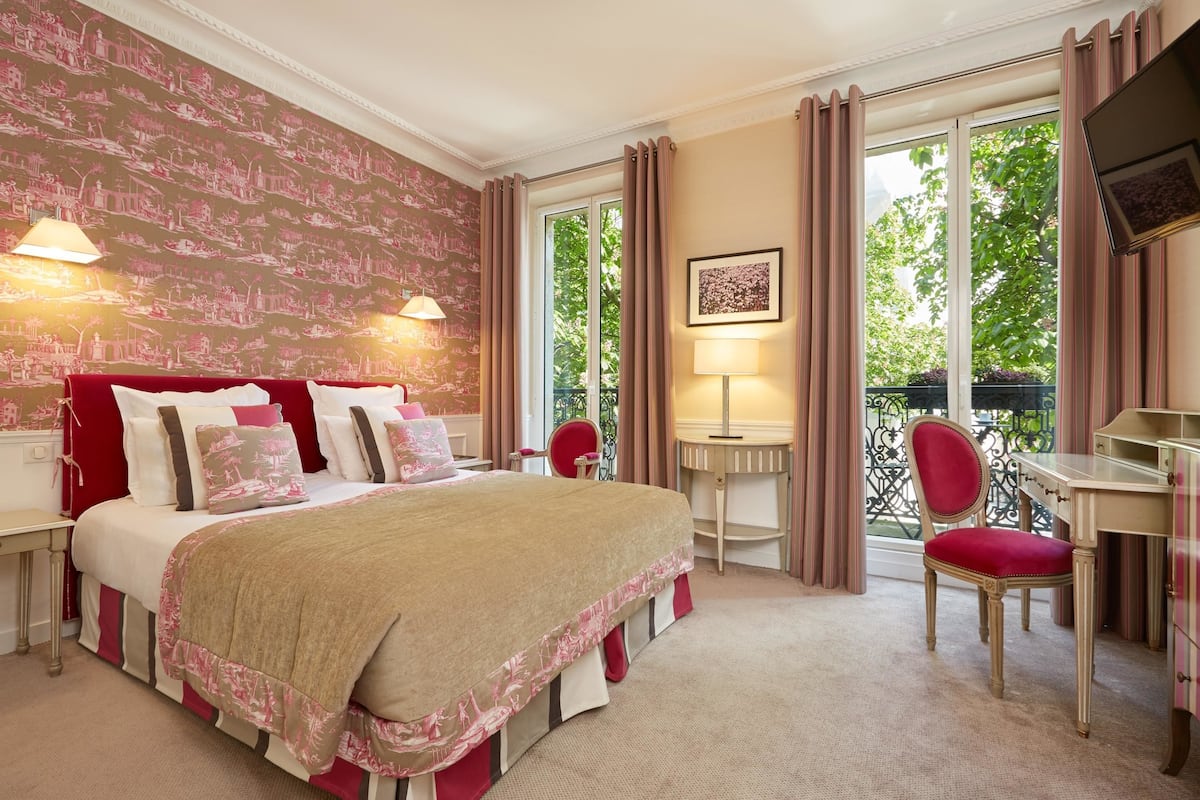 Habitación de hotel con una cama grande, un escritorio, una silla y un balcón con vistas a árboles.