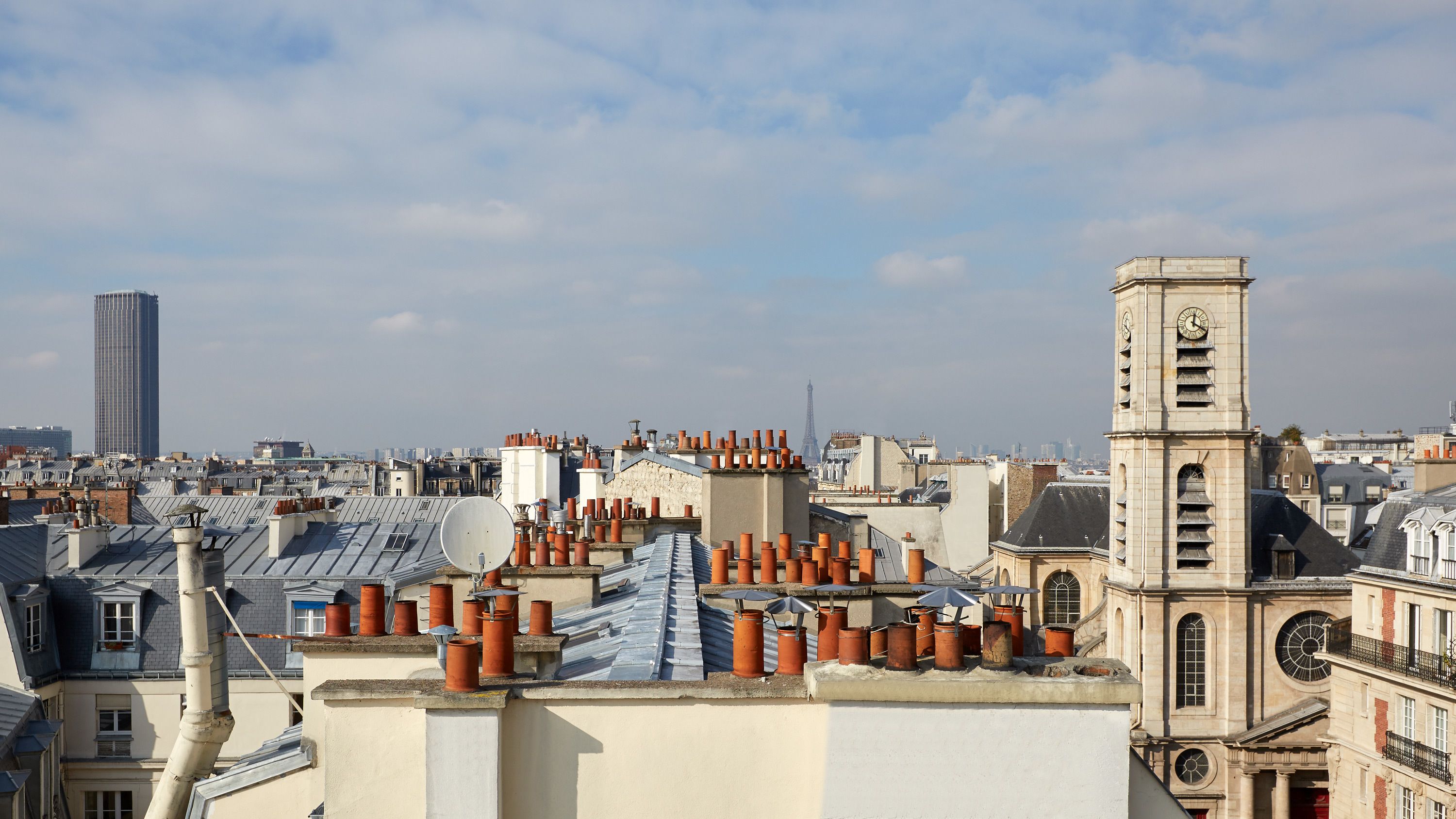 A cityscape with a prominent clock tower, a tall modern building, and numerous rooftops with satellite dishes and terracotta pots.