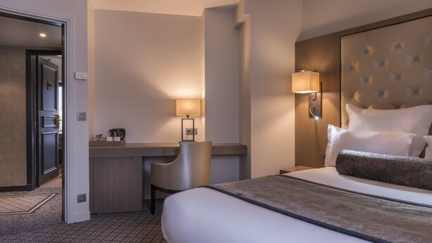 Habitación de hotel con cama, mesita de noche, lámpara, silla y vista al pasillo.