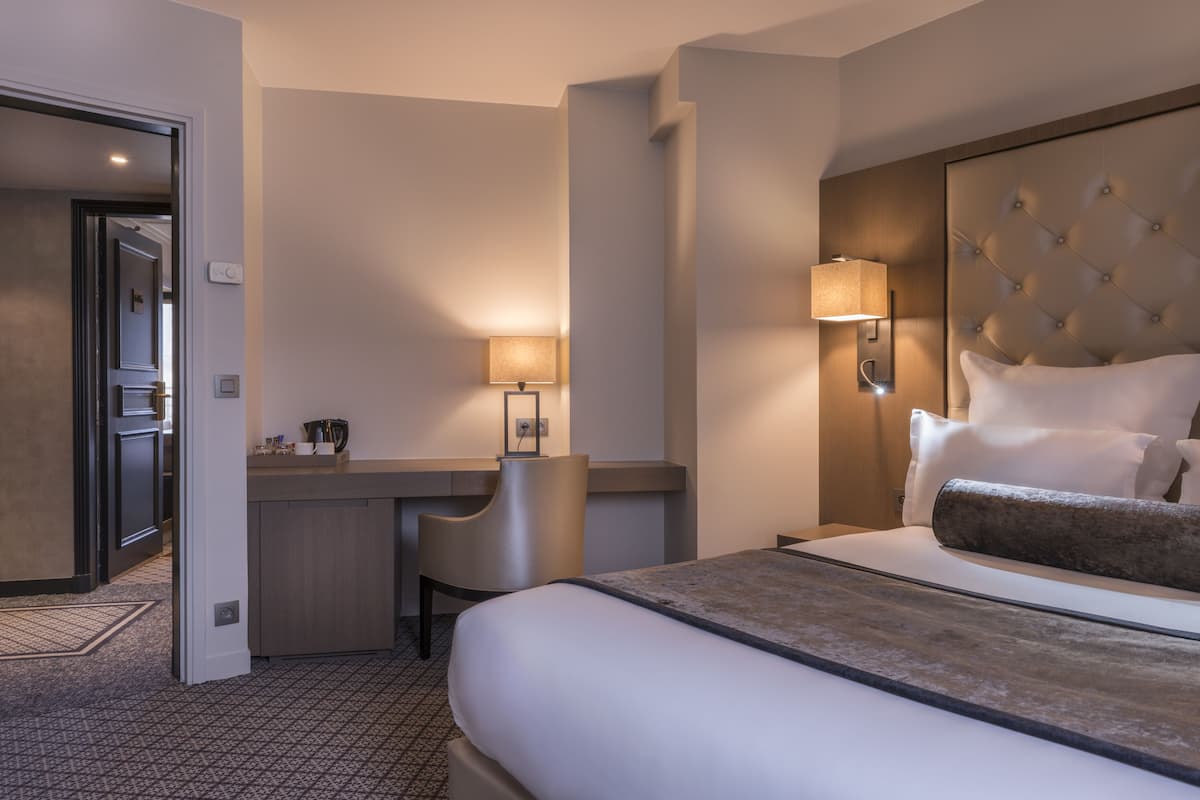 A hotel room with a bed, bedside table, lamp, chair, and a view of the hallway.