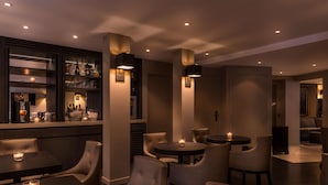 Zona de bar moderna con mesas redondas y sillones de cuero, una barra con botellas y vasos, e iluminación empotrada en el techo.