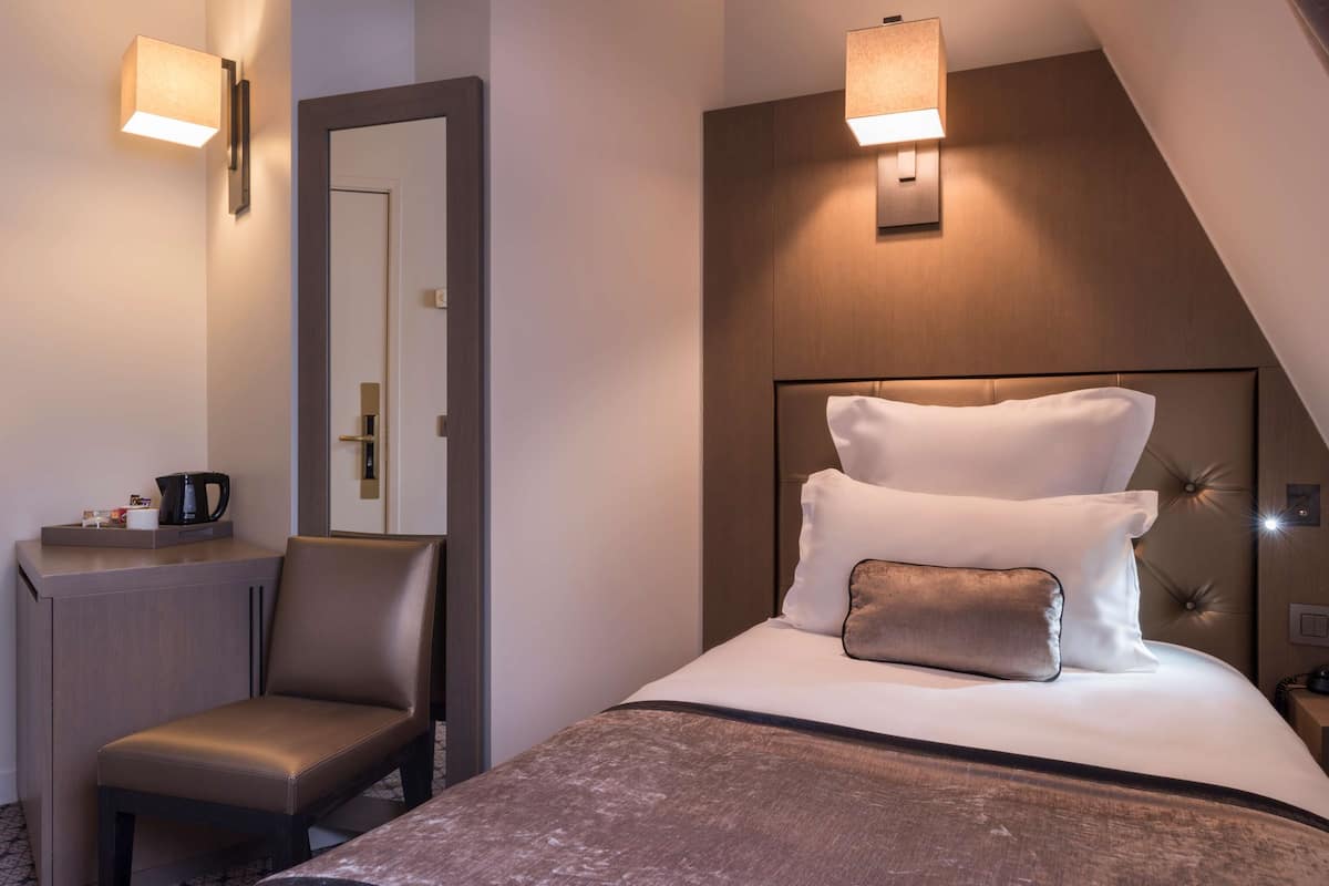 A modern hotel room with a bed, a desk, and a chair. There are wall-mounted lights above the bed and a mirror on the wall.