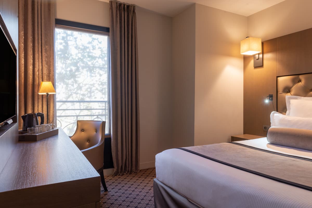 A hotel room with a bed, a desk with a lamp, a chair, and a window with curtains.