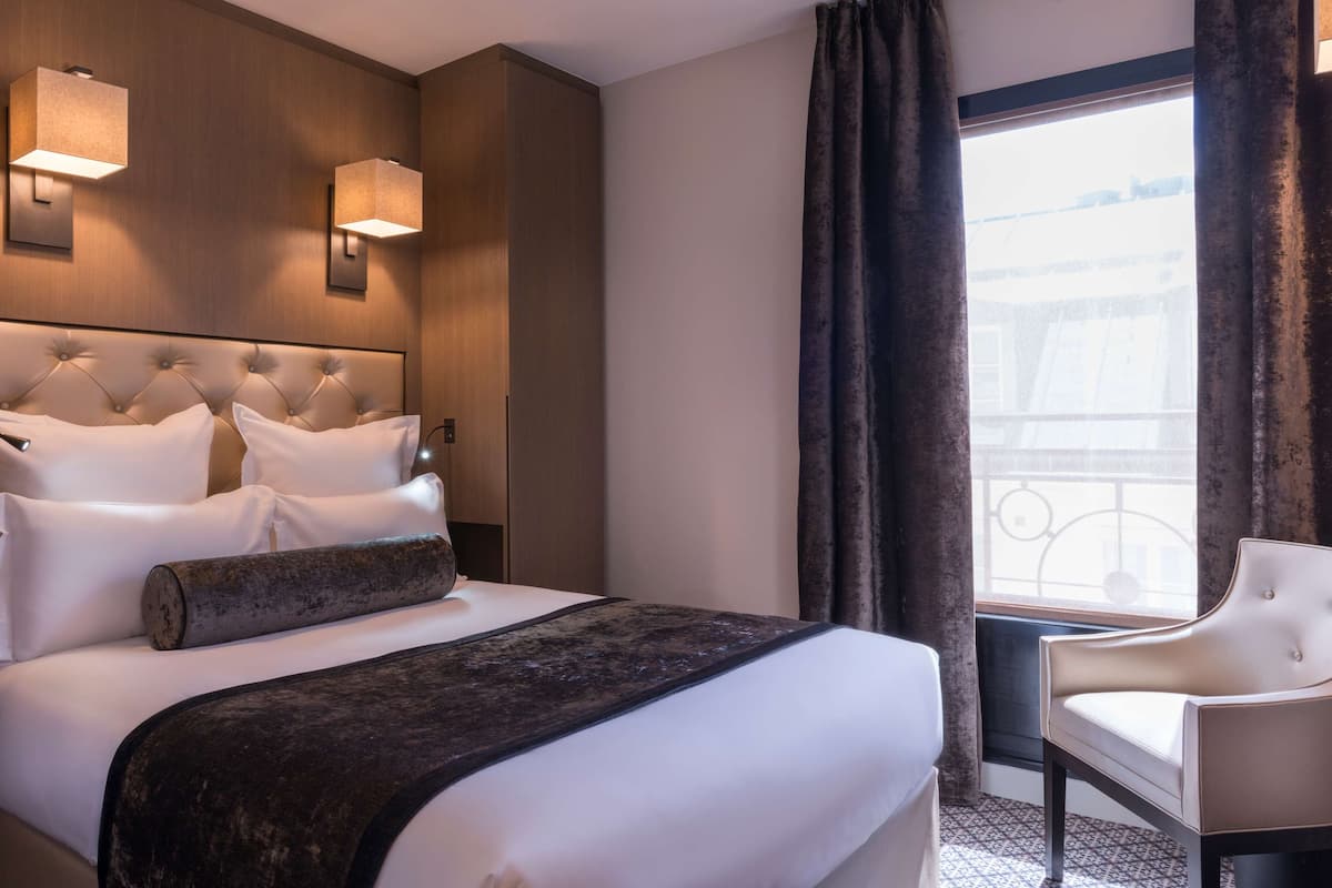 A hotel room with a large bed, a chair, a window with curtains, and wall-mounted lights.