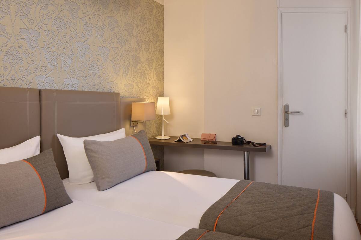 Une chambre d’hôtel avec un lit, des oreillers, une table de chevet avec une lampe, une commode et une porte.